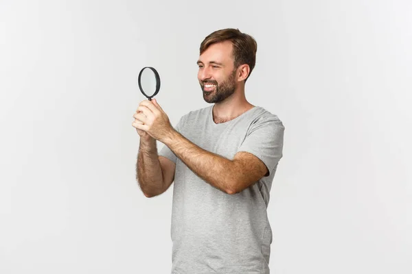 Bild av snygg kaukasisk man i grå t-shirt, söker efter något, ser nöjd genom förstoringsglas med nyfiken ansikte, står över vit bakgrund — Stockfoto