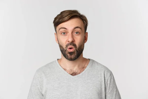 Κοντινό πλάνο ενός όμορφου ενήλικου άντρα με γενειάδα, με γκρι μπλουζάκι, που δείχνει έκπληκτος και περίεργος για τη διαφήμιση, να στέκεται πάνω από λευκό φόντο — Φωτογραφία Αρχείου