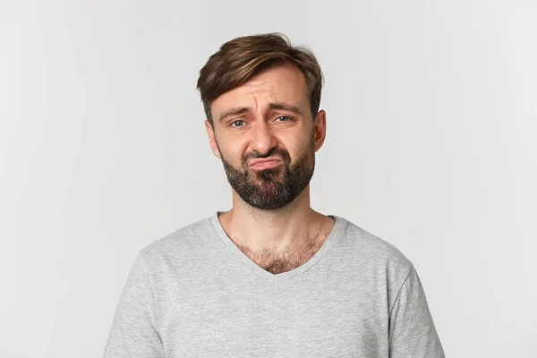 Close-up of gloomy disappointed guy with beard, wearing gray t-shirt, grimacing and frowning sad, standing over white background — Stock Photo, Image