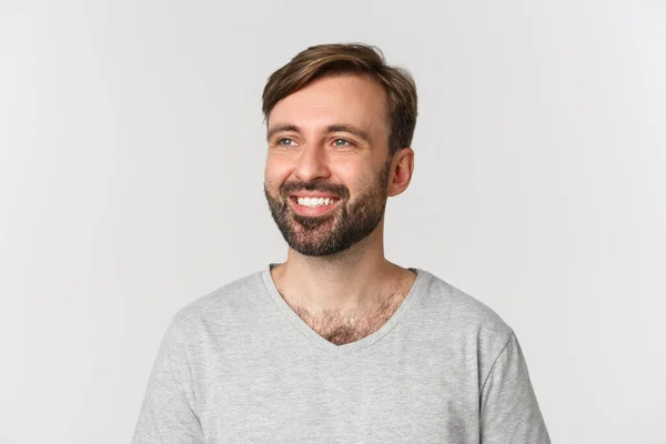 Retrato de homem barbudo bonito em camiseta cinza, olhando para a esquerda e sorrindo, em pé sobre fundo branco — Fotografia de Stock
