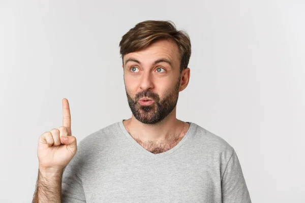 Close-up van doordachte man met baard in grijs t-shirt, met idee, vinger opsteken en omhoog kijken, over een witte achtergrond staan — Stockfoto