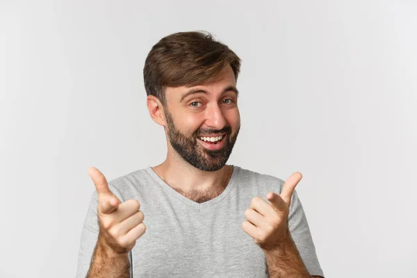 Close-up de homem adulto bonito com barba, sorrindo e apontando os dedos para a câmera, de pé em camiseta cinza sobre fundo branco — Fotografia de Stock