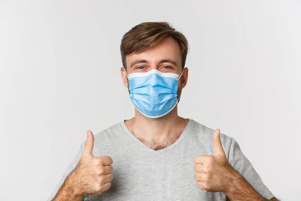 Concept of pandemic, covid-19 and social-distancing. Close-up of cheerful caucasian man in medical mask, showing thumbs-up, standing over white background — Stock Photo, Image