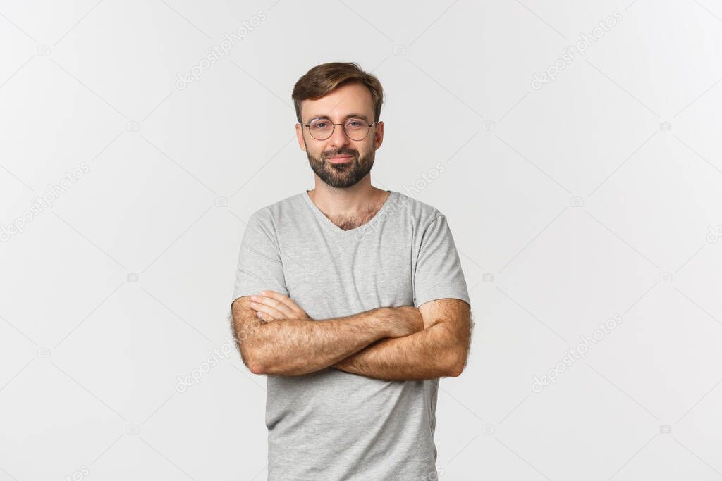 Image of confident handsome man in gray t-shirt and glasses, cross arms on chest and smiling, standing over white background