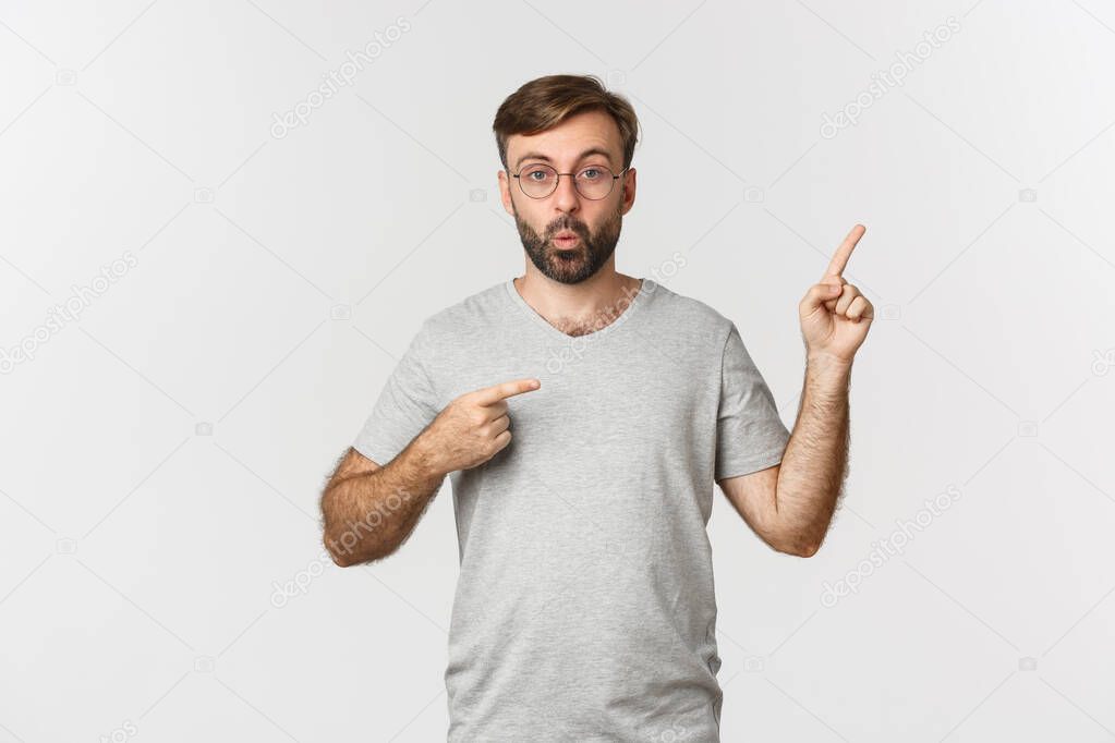 Amazed bearded man, saying wow, pointing fingers at upper right corner, showing logo, standing over white background