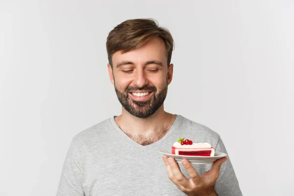 Close-up de homem barbudo bonito, sorrindo e olhando para bolo saboroso, de pé sobre fundo branco — Fotografia de Stock