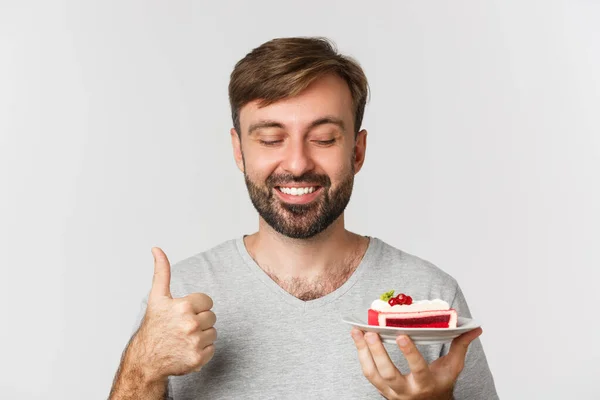 Close-up de cara caucasiano alegre com barba, vestindo camiseta cinza, segurando bolo e mostrando polegares para cima, como sobremesa, em pé sobre fundo branco — Fotografia de Stock