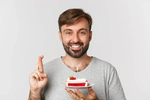 Close-up of hopeful man celebrating birthday, making wish with fingers crossed, holding bday cake, standing over white background — Stock Photo, Image
