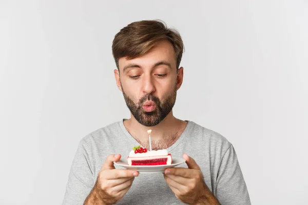 Close-up de homem barbudo feliz, sorrindo e comemorando aniversário, segurando bolo com vela acesa, fazendo desejo b-dia, de pé sobre fundo branco — Fotografia de Stock