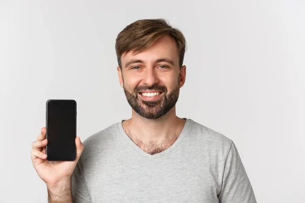Крупный план улыбающегося красавца в серой футболке, показывающего экран мобильного телефона, рекомендующего приложение или торговый сайт, стоящего на белом фоне — стоковое фото