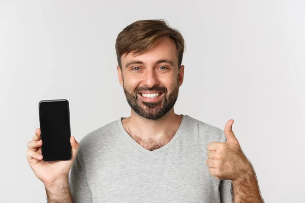 Närbild av leende stilig man i grå t-shirt, visar mobiltelefon skärm och tummen upp, rekommendera app, står över vit bakgrund — Stockfoto