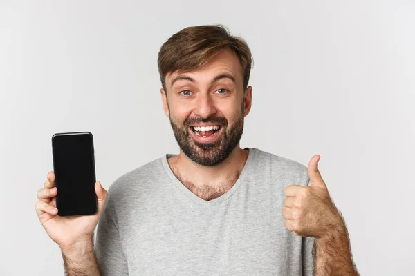 Крупный план улыбающегося красавца в серой футболке, показывающего экран мобильного телефона и большой палец вверх, рекомендующего приложение, стоящего на белом фоне — стоковое фото