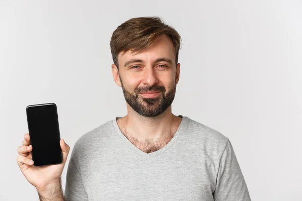 Närbild av nöjd leende man med skägg, ser glad och visar mobiltelefon skärm, står över vit bakgrund — Stockfoto