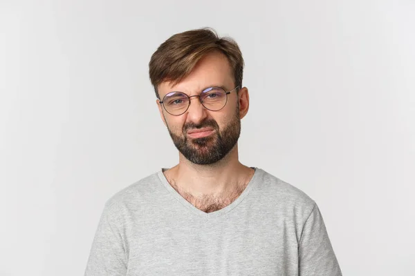 Close-up de homem barbudo cético em óculos e camiseta cinza, sorridente decepcionado, de pé sobre fundo branco — Fotografia de Stock