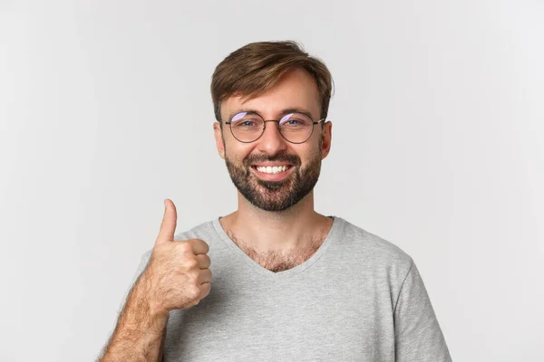 Primer plano del alegre hombre caucásico con barba, con gafas y camiseta casual, sonriendo y mostrando los pulgares hacia arriba en aprobación, recomiendan el producto, de pie sobre fondo blanco —  Fotos de Stock