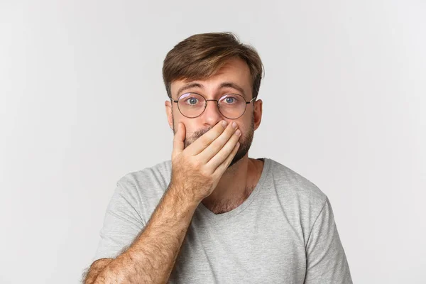 Close-up of shocked and worried man in glasses, gasping and cover mouth, hear something scary, standing over white background — Stock Photo, Image