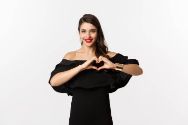 Moda ve güzellik. Güzel, genç bir kadın kalp işareti ve gülümseme gösteriyor, sevgisini ve sempatisini ifade ediyor, siyah elbise giyiyor, beyaz arka plan