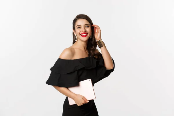 Concepto de moda y compras. Mujer joven con estilo con maquillaje glamour, vestido negro, la celebración de la tableta digital y sonriente, fondo blanco — Foto de Stock