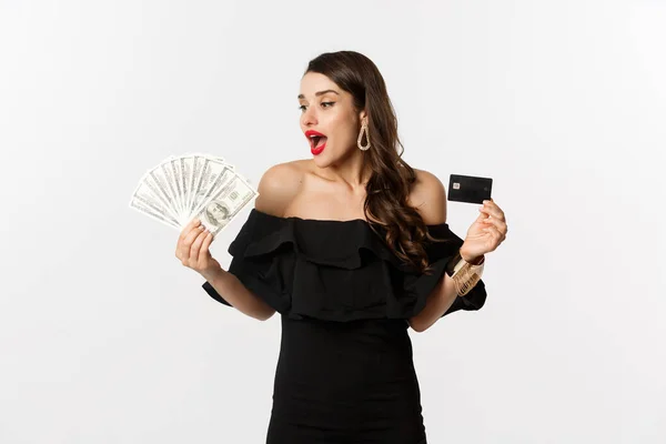 Schoonheid en winkelen concept. Fashionable vrouw met credit card, kijken naar geld met verbazing, staande over witte achtergrond — Stockfoto