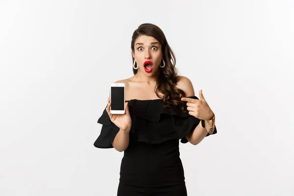 Nätköpskoncept. Fashionabel kvinna i svart klänning pekar finger på smartphone skärm, visar ansökan, står över vit bakgrund — Stockfoto