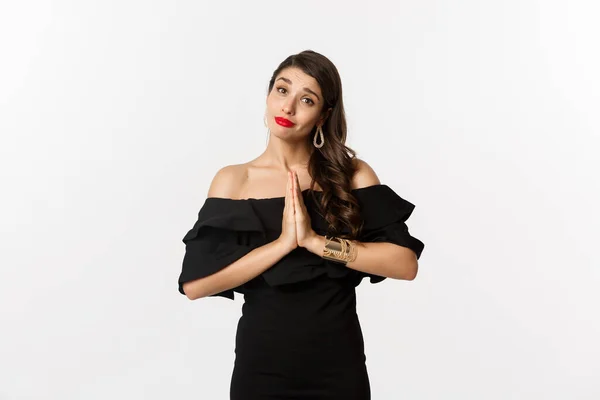 Moda y belleza. Mujer bastante elegante pidiendo ayuda, rogando por algo, decir por favor con las manos juntas, de pie en vestido negro, fondo blanco — Foto de Stock