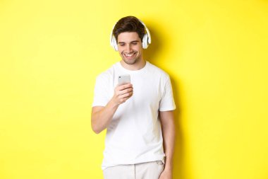 Kulaklıklı genç adam akıllı telefondan mesaj okuyor, gülümsüyor, kulaklıkla müzik dinliyor ve uygulamadan şarkı seçiyor, sarı arka planda duruyor.