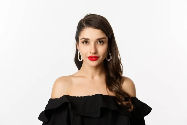 Conceito de moda e beleza. Close-up de mulher morena elegante com brincos, vestindo vestido preto e batom vermelho, olhando sensual para a câmera, em pé sobre fundo branco — Fotografia de Stock