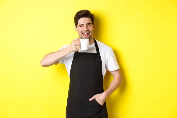 Bonito barista bebendo xícara de café e piscando, convidando para café, de pé sobre fundo amarelo — Fotografia de Stock