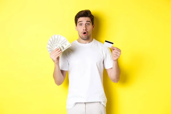 Hombre sorprendido sosteniendo tarjeta de crédito y dinero, mirando sorprendido, de pie sobre fondo amarillo — Foto de Stock