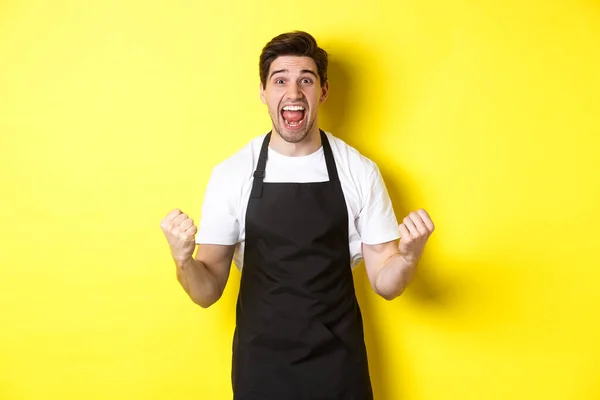Ενθουσιασμένος ιδιοκτήτης καφετέριας με μαύρη ποδιά γιορτάζει, κάνοντας αντλία γροθιά και φωνάζοντας για τη χαρά, επιτύχει το στόχο, στέκεται ενάντια στο κίτρινο φόντο — Φωτογραφία Αρχείου