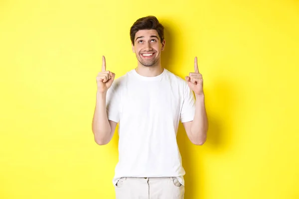 Excited attractive man in white t-shirt, pointing fingers up, looking at advertisement with happy smile, standing over yellow background — Stock Photo, Image