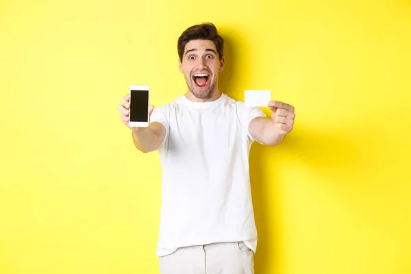 stock image Handsome caucasian man showing smartphone screen and credit card, concept of mobile banking and online shopping, yellow background