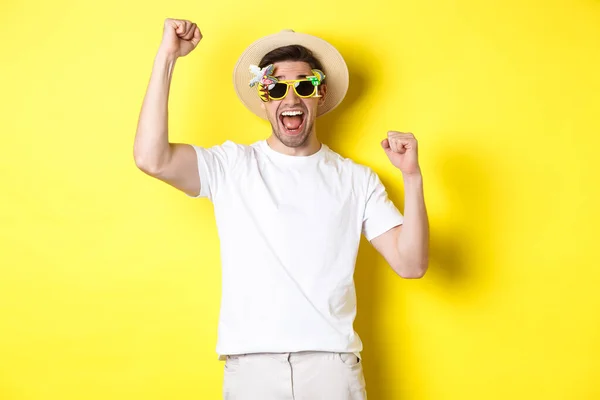 Concepto de turismo y estilo de vida. Feliz hombre ganando viaje al resort, gritando sí y levantando las manos, triunfando, usando gafas de sol y sombrero de verano, fondo amarillo — Foto de Stock