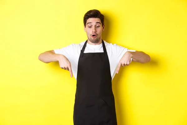 Surpreendido barista masculino em avental preto verificando oferta promocional, apontando os dedos para baixo no banner logotipo, de pé sobre fundo amarelo — Fotografia de Stock