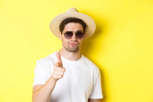 Concepto de turismo y vacaciones. Primer plano del chico fresco en sombrero de verano y gafas de sol apuntando con el dedo a la cámara, de pie sobre el fondo amarillo — Foto de Stock