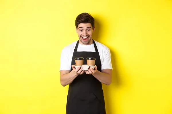 Camarero mirando emocionado a dos tazas de café para llevar, con delantal negro, de pie sobre el fondo amarillo — Foto de Stock