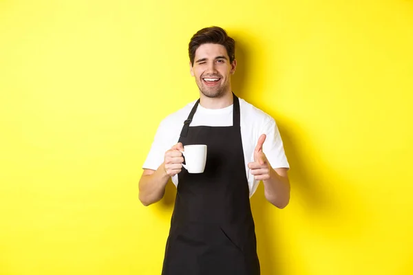 Barista tampan dengan celemek hitam memegang cangkir kopi, menunjuk jari padamu, mengundang kafe, berdiri di atas latar belakang kuning — Stok Foto