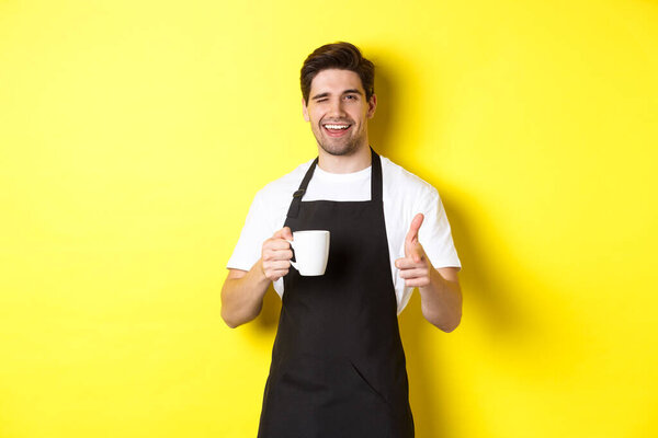 Handsome barista in black apron holding coffee cup, pointing finger at you, inviting visit cafe, standing over yellow background