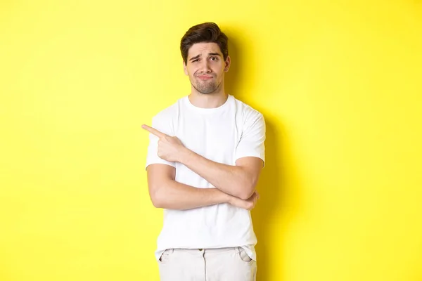 Скептически настроенный и неохотный парень в белой футболке, указывающий пальцем влево и ухмыляющийся незабавно, показывающий плохую рекламу, стоящий на желтом фоне — стоковое фото
