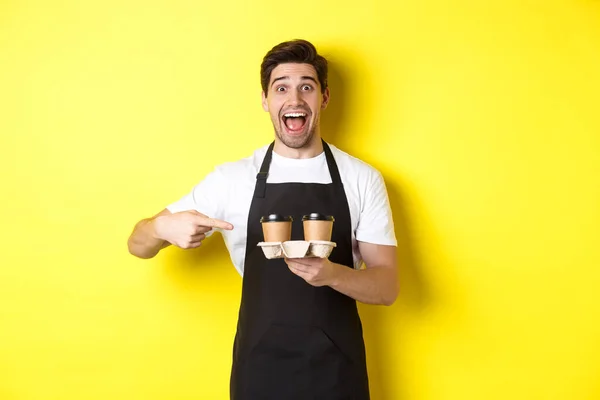 Захоплений бариста в чорному фартусі, спрямований пальцями на винос кавових чашок, стоїть на жовтому фоні щасливий — стокове фото