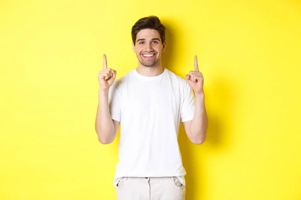 Handsome guy in white t-shirt pointing fingers up, showing shopping offers, standing over yellow background — Stock Photo, Image