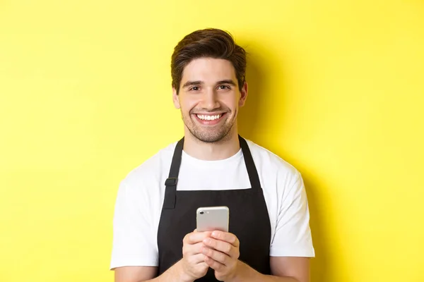 Крупним планом красивий бариста надсилає повідомлення на мобільний телефон, посміхаючись щасливим, стоїть над жовтим тлом — стокове фото