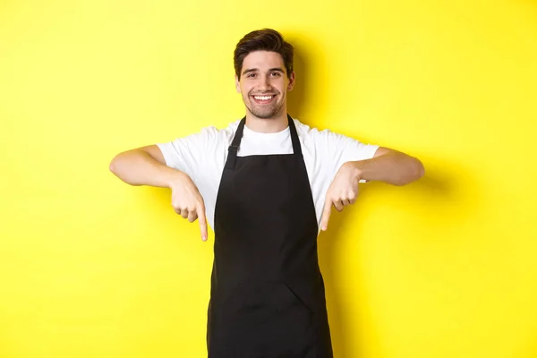 Vriendelijke barista in zwarte schort wijzende vingers naar beneden, met uw logo banner, staande over gele achtergrond — Stockfoto