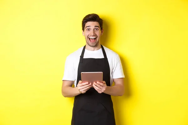 Χαρούμενος πωλητής με μαύρη ποδιά, κρατώντας ψηφιακό tablet και δείχνοντας έκπληκτος, στέκεται απέναντι σε κίτρινο φόντο — Φωτογραφία Αρχείου