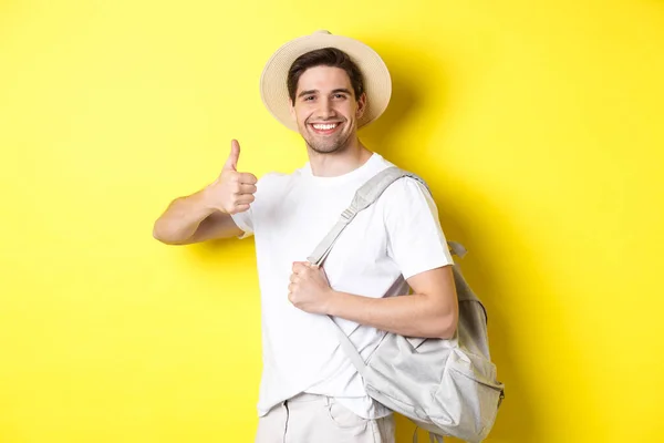 Concepto de turismo, viajes y vacaciones. Hombre feliz yendo de vacaciones, sosteniendo la mochila y mostrando el pulgar hacia arriba, sonriendo satisfecho, de pie sobre fondo amarillo — Foto de Stock