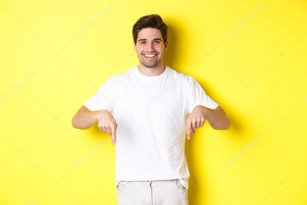 Attractive bearded man pointing fingers down, showing event banner, standing over yellow background.