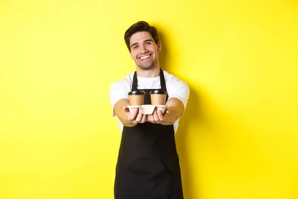 Amistoso barista en delantal negro dando orden de comida para llevar, sosteniendo dos tazas de café y sonriendo, de pie sobre fondo amarillo —  Fotos de Stock