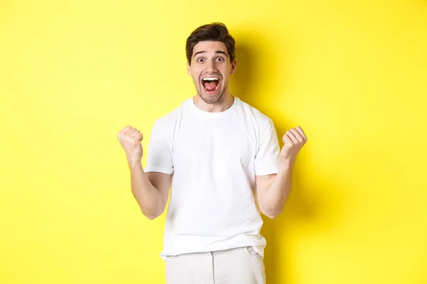 Hombre excitado y afortunado ganando, apretando puños y luciendo feliz, triunfando y celebrando, de pie sobre fondo amarillo —  Fotos de Stock