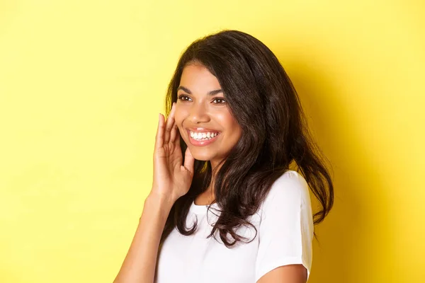 Close-up van mooie Afrikaans-Amerikaanse vrouw, glimlachend en kijkend naar links, zacht aandoend gezicht na het aanbrengen van make-up product, staande over gele achtergrond — Stockfoto