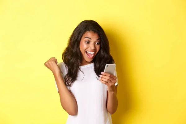 Retrato de la hermosa chica afroamericana, regocijándose y mirando el teléfono móvil, encontró algo increíble en línea, de pie sobre el fondo amarillo — Foto de Stock